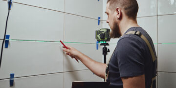 Close-up of a person regrouting bathroom tiles using a grout float, removing old grout and applying fresh sealant between white ceramic tiles.