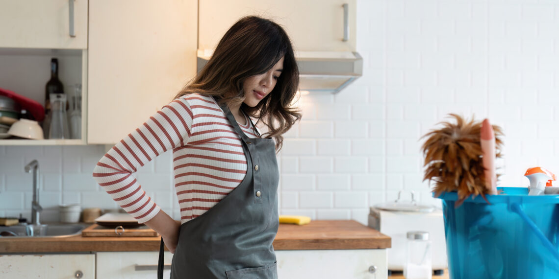 Woman in a kitchen wearing an apron and holding her lower back while preparing to start cleaning, with a bucket of supplies nearby.