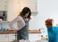 Woman in a kitchen wearing an apron and holding her lower back while preparing to start cleaning, with a bucket of supplies nearby.