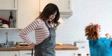 Woman in a kitchen wearing an apron and holding her lower back while preparing to start cleaning, with a bucket of supplies nearby.