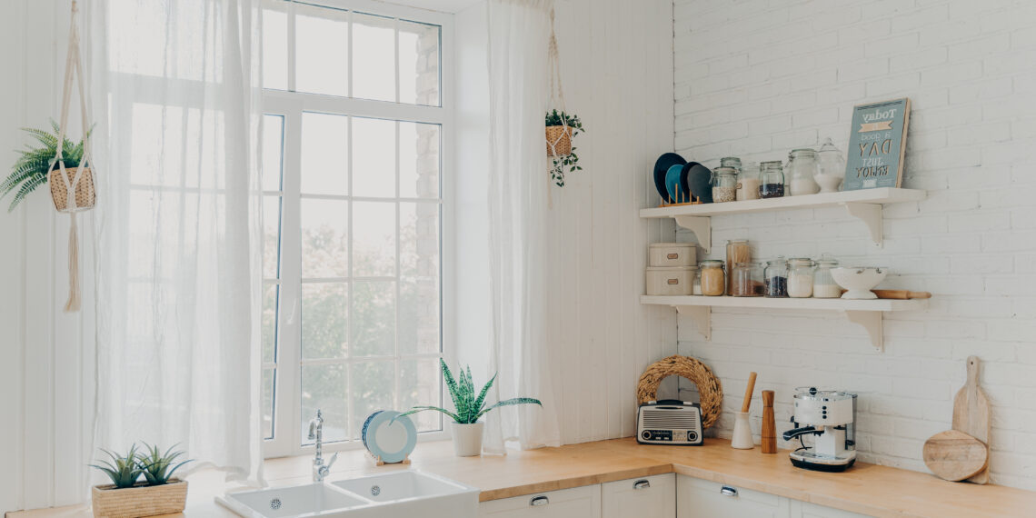 Bright budget-friendly kitchen with white cabinets, rustic wooden worktops, open shelving, and natural light streaming through large windows.