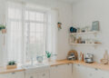 Bright budget-friendly kitchen with white cabinets, rustic wooden worktops, open shelving, and natural light streaming through large windows.
