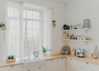 Bright budget-friendly kitchen with white cabinets, rustic wooden worktops, open shelving, and natural light streaming through large windows.