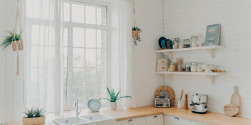 Bright budget-friendly kitchen with white cabinets, rustic wooden worktops, open shelving, and natural light streaming through large windows.