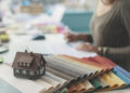 Interior design workspace featuring fabric swatches, a miniature house model, and a designer reviewing home extension or renovation plans.
