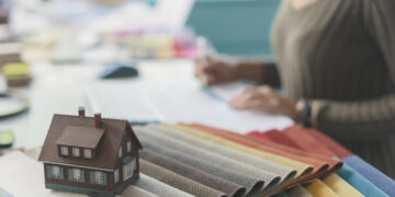 Interior design workspace featuring fabric swatches, a miniature house model, and a designer reviewing home extension or renovation plans.