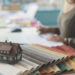 Interior design workspace featuring fabric swatches, a miniature house model, and a designer reviewing home extension or renovation plans.