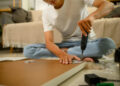 A person using an electric screwdriver to assemble flat pack furniture while sitting on the floor surrounded by wooden panels, screws, and packaging in a cosy living room.