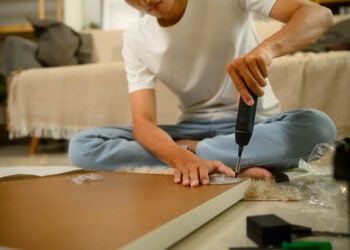 A person using an electric screwdriver to assemble flat pack furniture while sitting on the floor surrounded by wooden panels, screws, and packaging in a cosy living room.