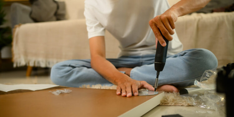 A person using an electric screwdriver to assemble flat pack furniture while sitting on the floor surrounded by wooden panels, screws, and packaging in a cosy living room.