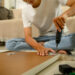 A person using an electric screwdriver to assemble flat pack furniture while sitting on the floor surrounded by wooden panels, screws, and packaging in a cosy living room.