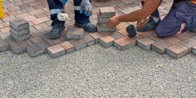 Workers installing a block paving driveway by carefully laying brick pavers over a gravel base outdoors