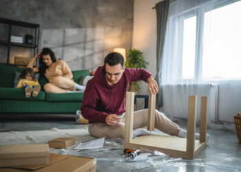 A man assembling flat pack furniture on the floor of a modern living room while his family relaxes in the background, surrounded by boxes, tools, and instruction sheets.
