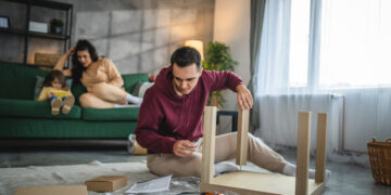 A man assembling flat pack furniture on the floor of a modern living room while his family relaxes in the background, surrounded by boxes, tools, and instruction sheets.
