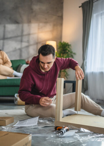 A man assembling flat pack furniture on the floor of a modern living room while his family relaxes in the background, surrounded by boxes, tools, and instruction sheets.